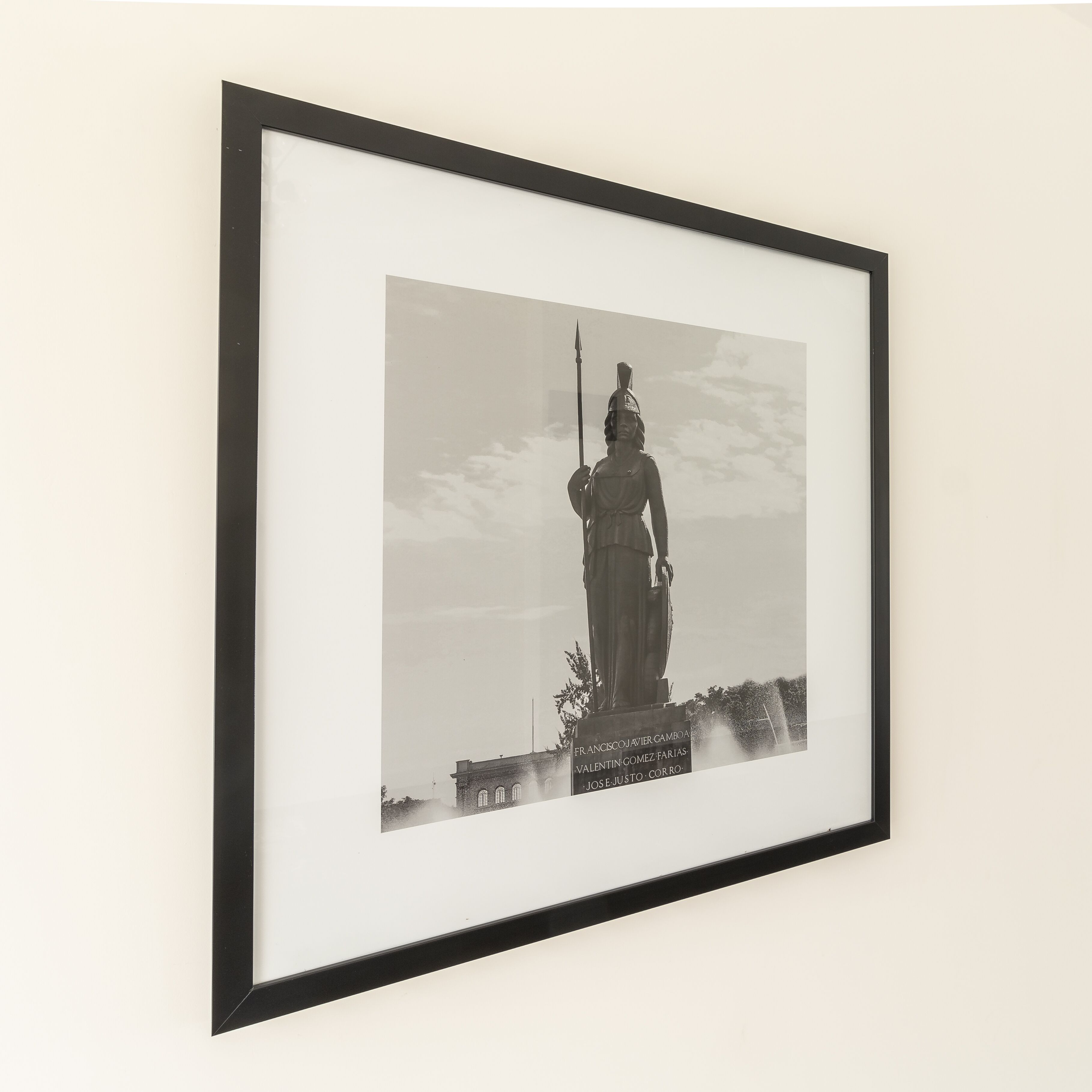 Een zwart-wit foto in een lijst van een standbeeld met een voetstuk waarop een inscriptie staat.