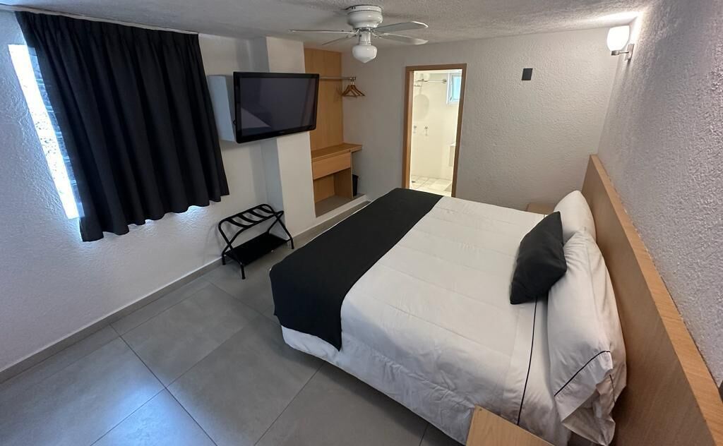 A hotel room with a bed, a TV mounted on the wall, a ceiling fan, and a window with curtains.