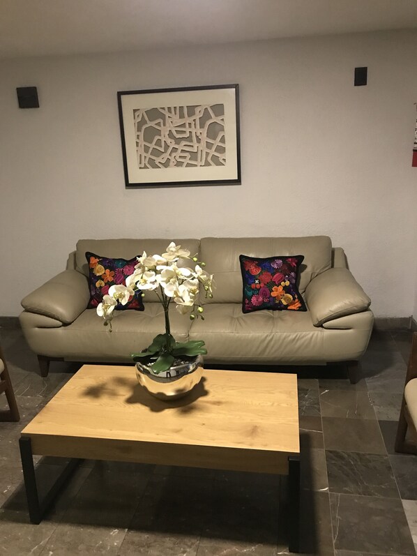 A living room with a beige sofa, two colorful pillows, a wooden coffee table, and a framed abstract artwork on the wall.