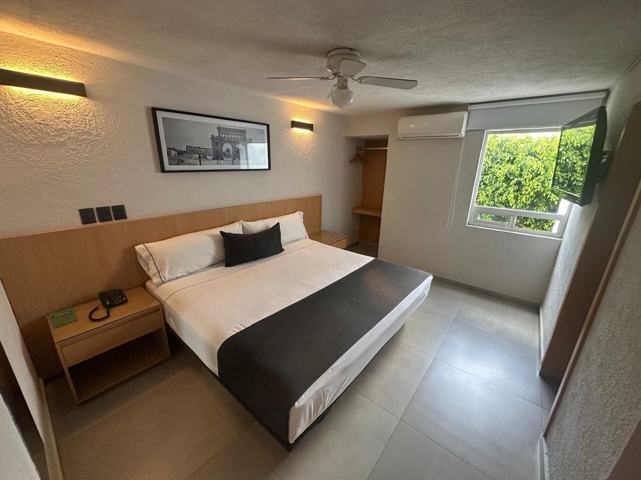 A hotel room with a bed, bedside table, nightstand, wall-mounted phone, framed picture, ceiling fan, window with view, and built-in air conditioning unit.
