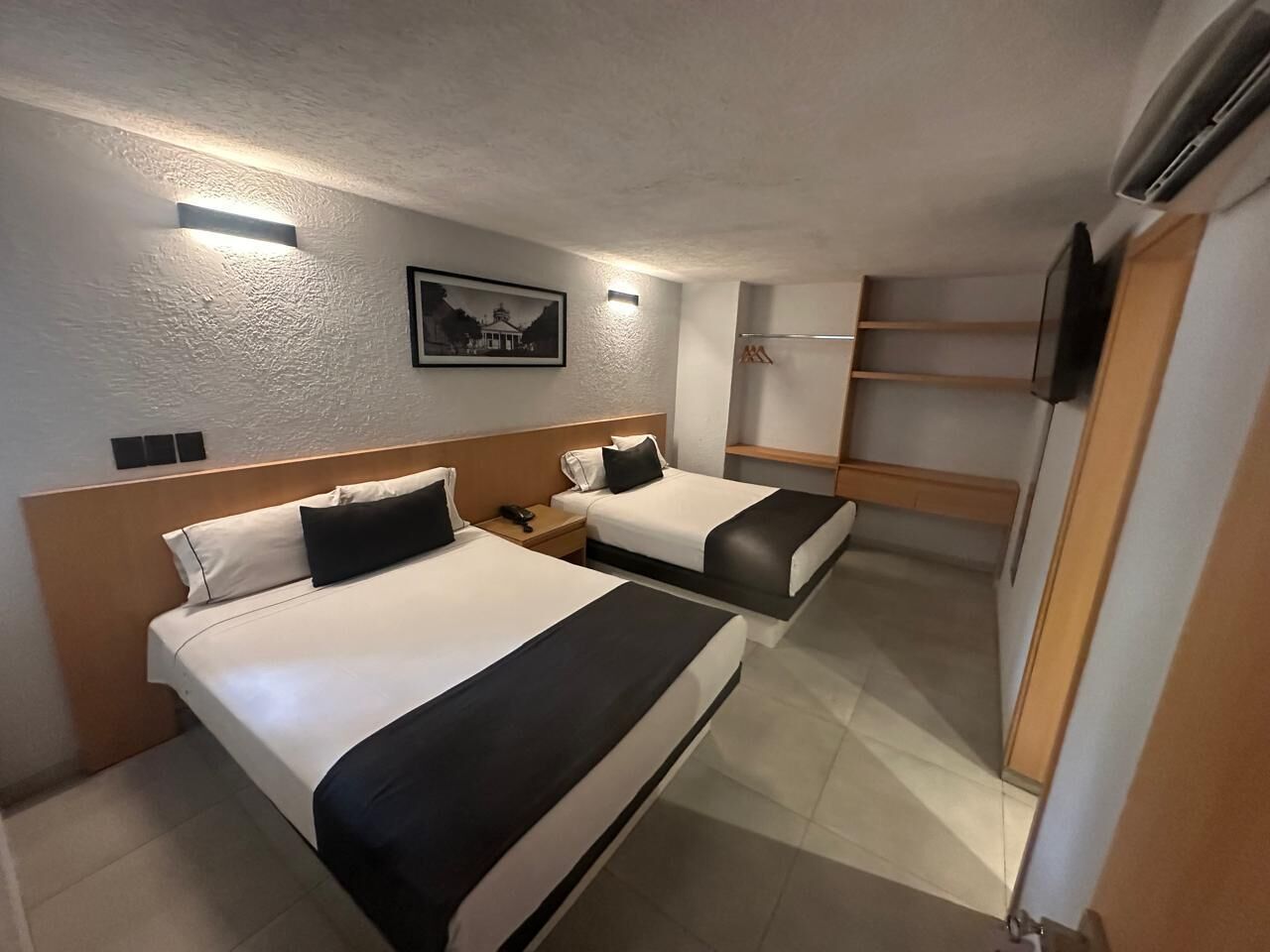 Habitació d'hotel amb dos llits, una tauleta de nit, un televisor i una estant penjada a la paret.