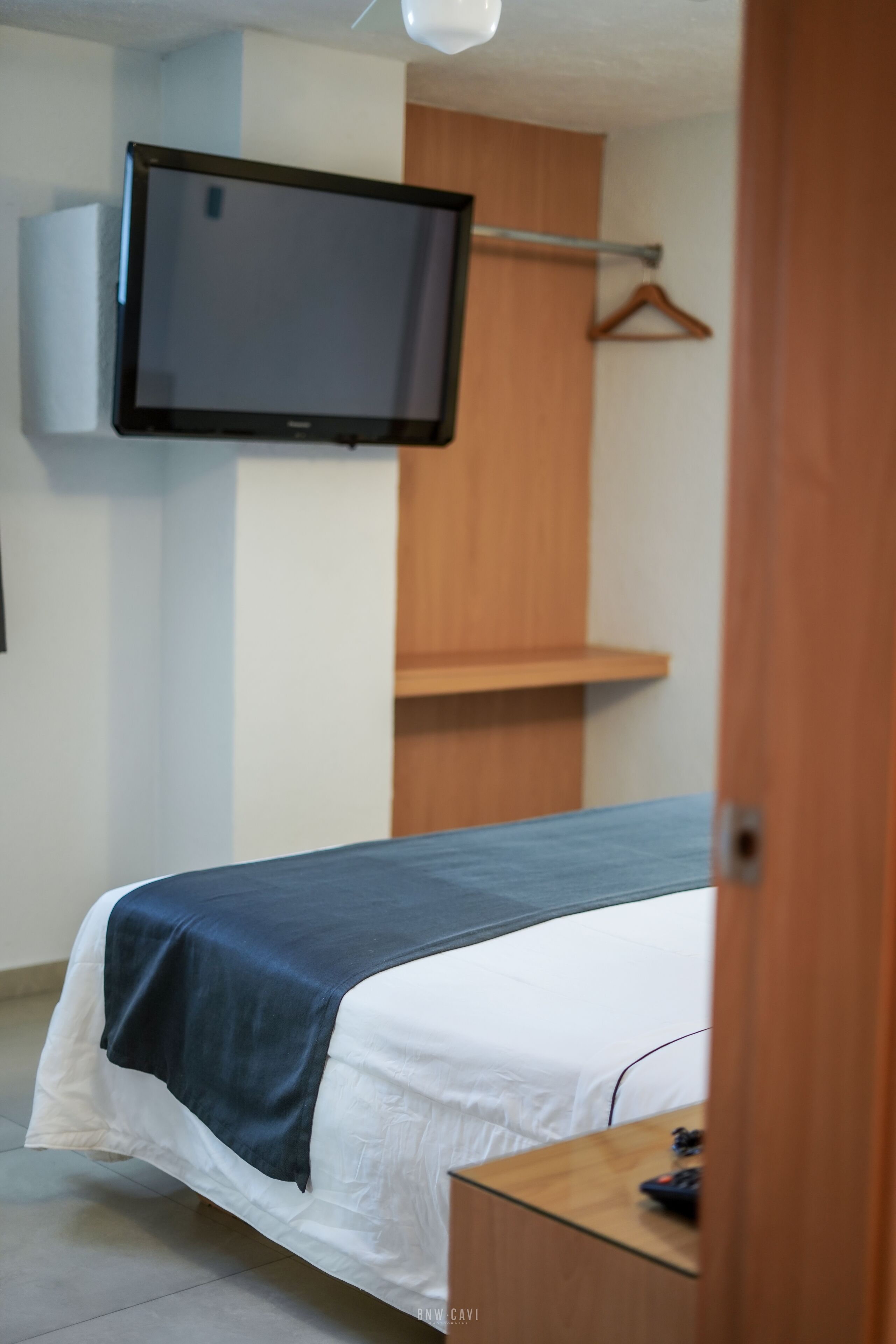 A bedroom with a bed, a TV mounted on the wall, a wooden wardrobe, and a small table with a remote control.