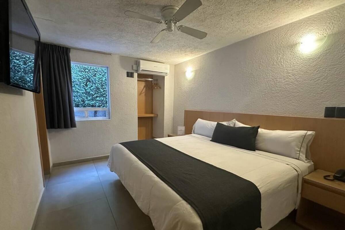 Una habitación de hotel con cama, ventilador de techo, televisión, ventana con cortinas y una pequeña repisa.
