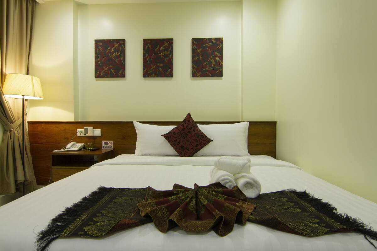 A hotel room with a neatly made bed, a nightstand with a phone and a "No Smoking" sign, and three abstract paintings on the wall.