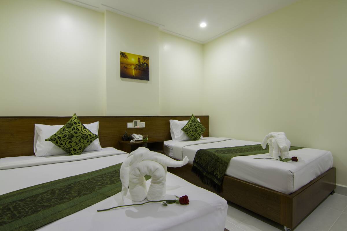 A hotel room with three beds, each with a green bedspread and a white towel shaped like an elephant. There is a painting on the wall and a wooden headboard.