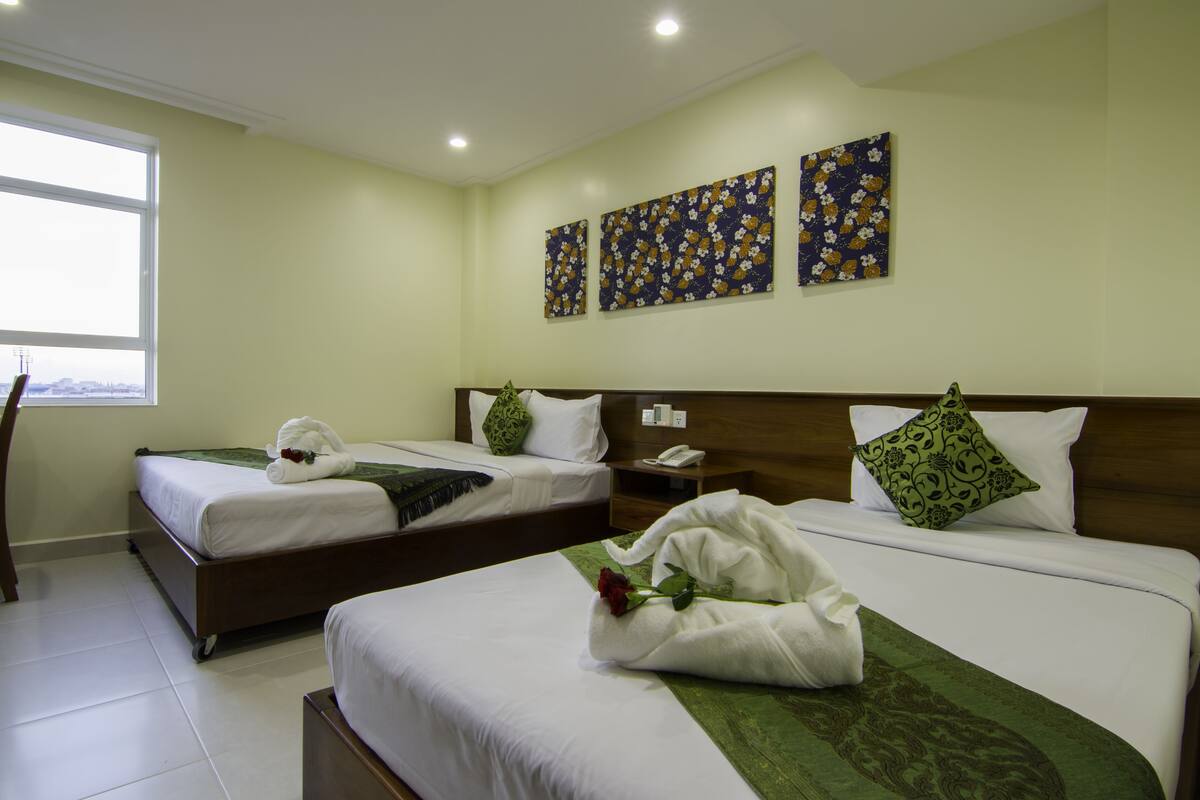 A hotel room with two beds, each with white linens and green decorative pillows. There are three floral artwork pieces on the wall, and a window providing a view.