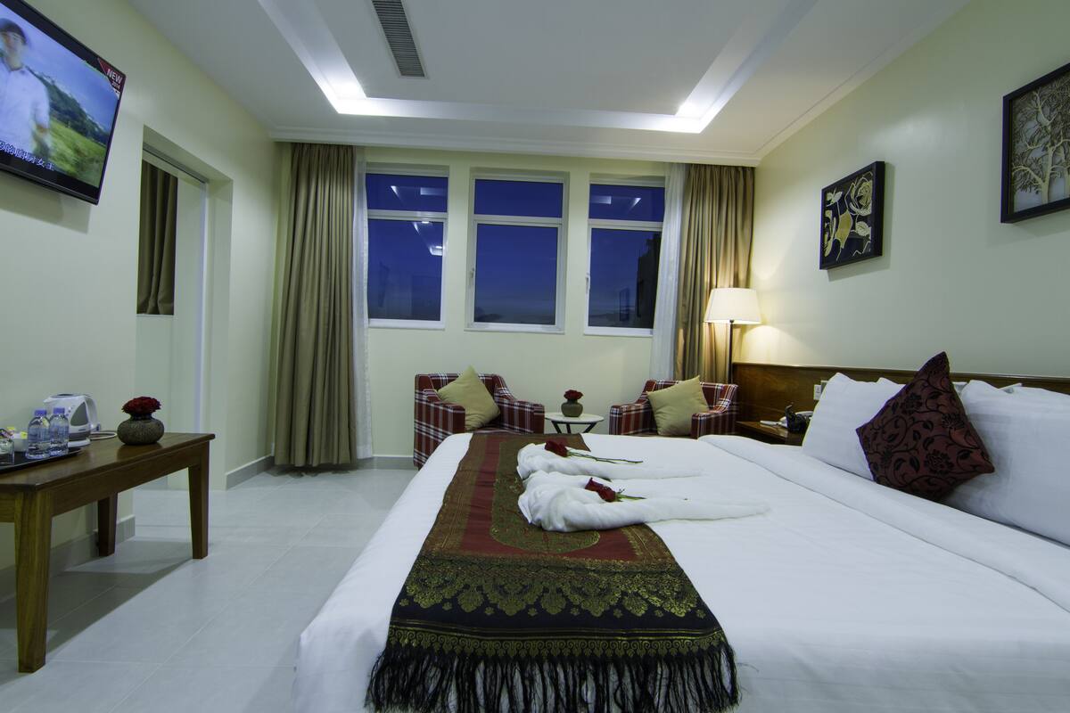 A hotel room with a large bed, a TV, a table with a vase and a flower, and a window with curtains.
