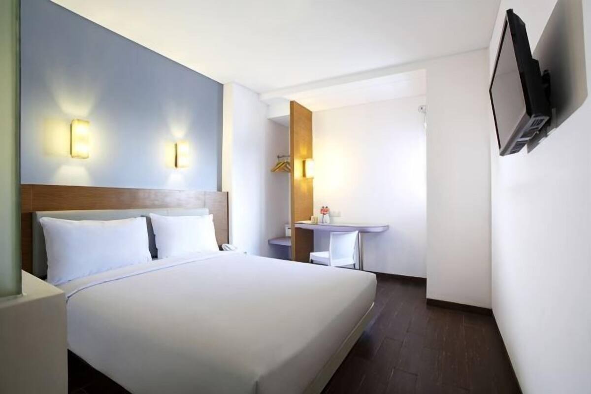 A hotel room with a bed, bedside tables, a desk, a chair, and a TV mounted on the wall.