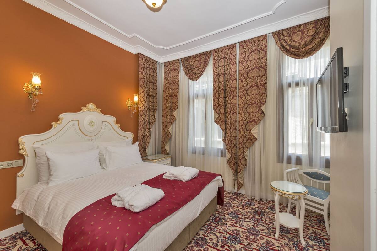 A hotel room with a large bed, a small table, a TV, and ornate curtains.