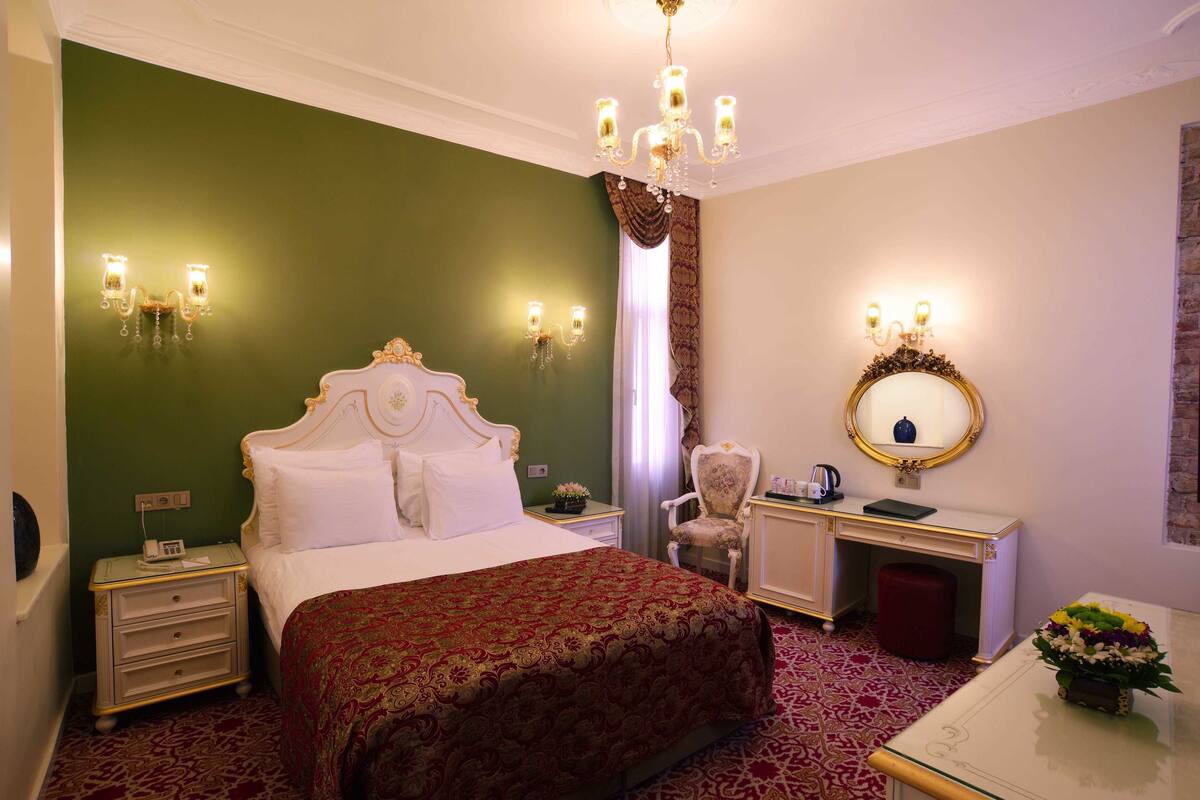 A hotel room with a large bed, a desk, a chair, a vanity with a mirror, and a chandelier.