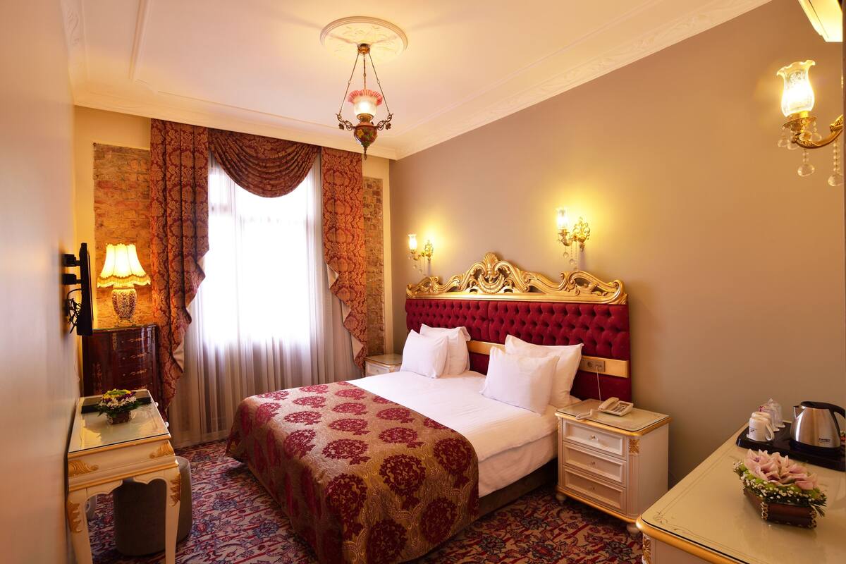 A hotel room with a large bed, a desk, a chair, a nightstand, a lamp, a chandelier, and a window with curtains.