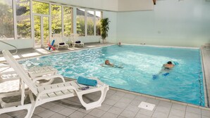 An indoor swimming pool with clear blue water, surrounded by white lounge chairs and a large window offering a view of greenery outside.