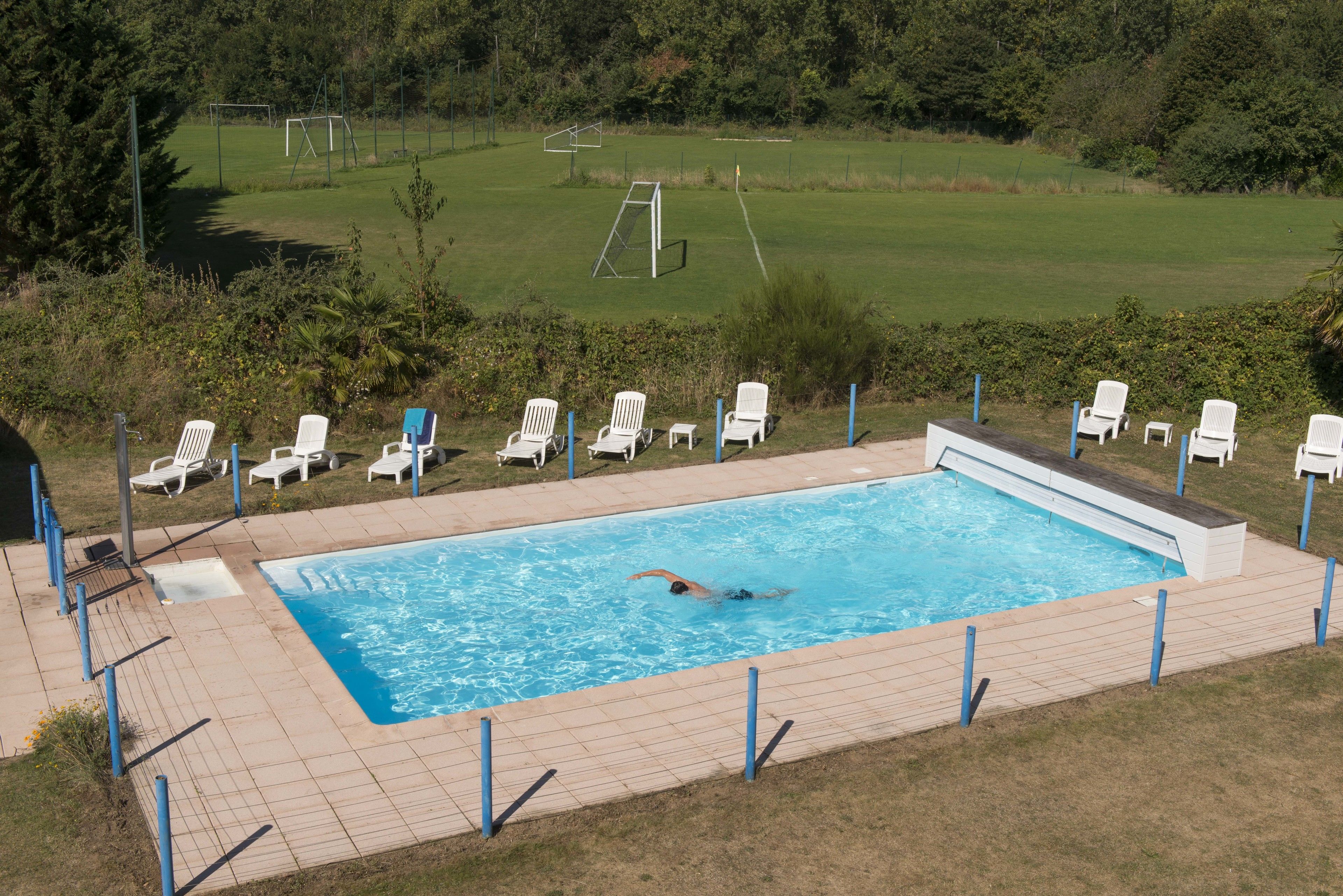 Una piscina con una persona nadando, rodeada de sillas de descanso blancas y un césped con una portería de fútbol al fondo.