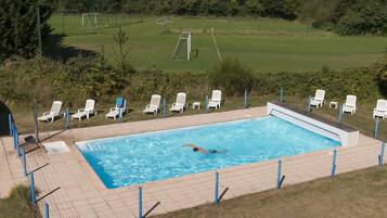 Una piscina con una persona nadando, rodeada de sillas de descanso blancas y un césped con una portería de fútbol al fondo.