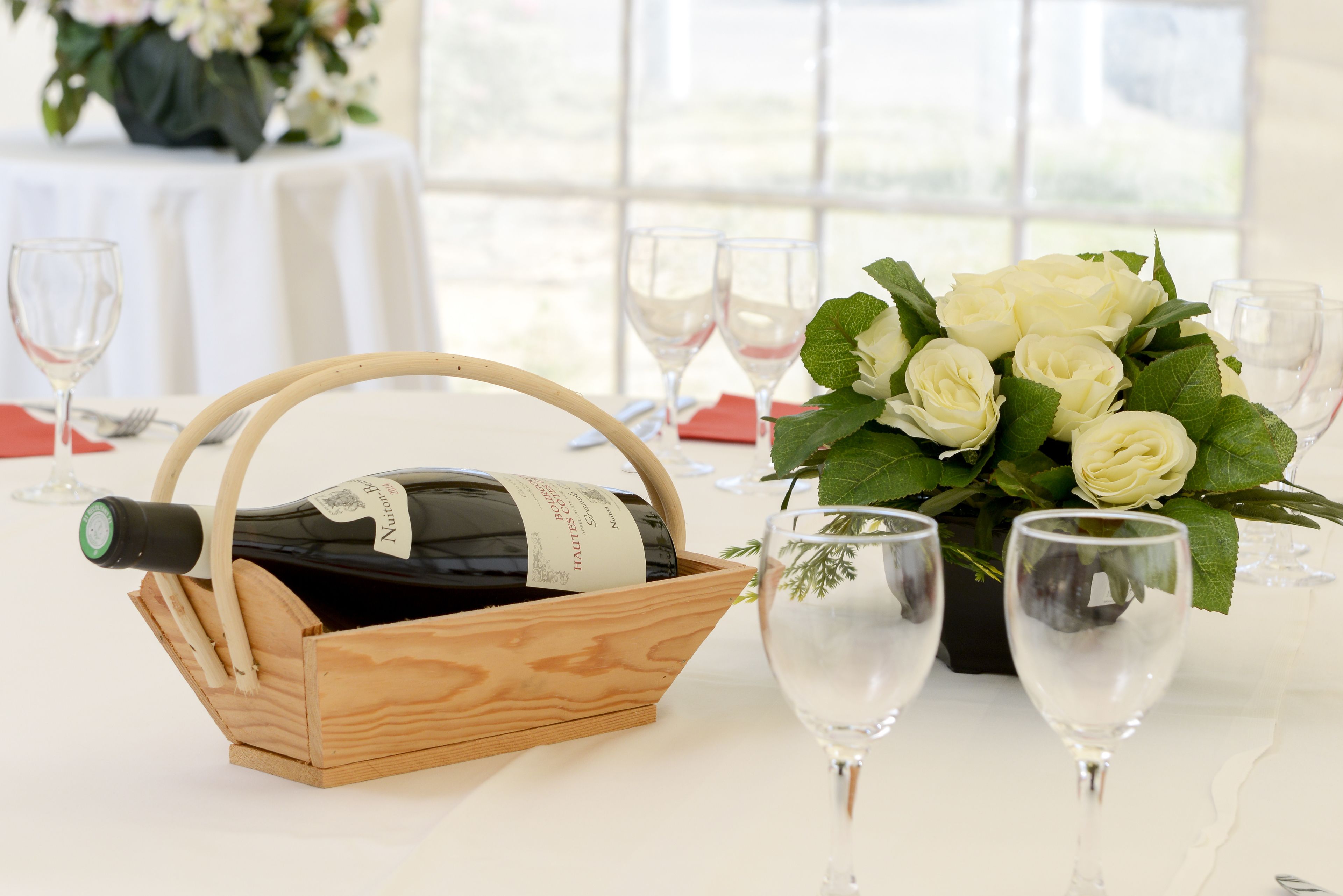 Een gedekte tafel met een wijnfles in een houten mand, een boeket witte rozen en verschillende wijnglazen.