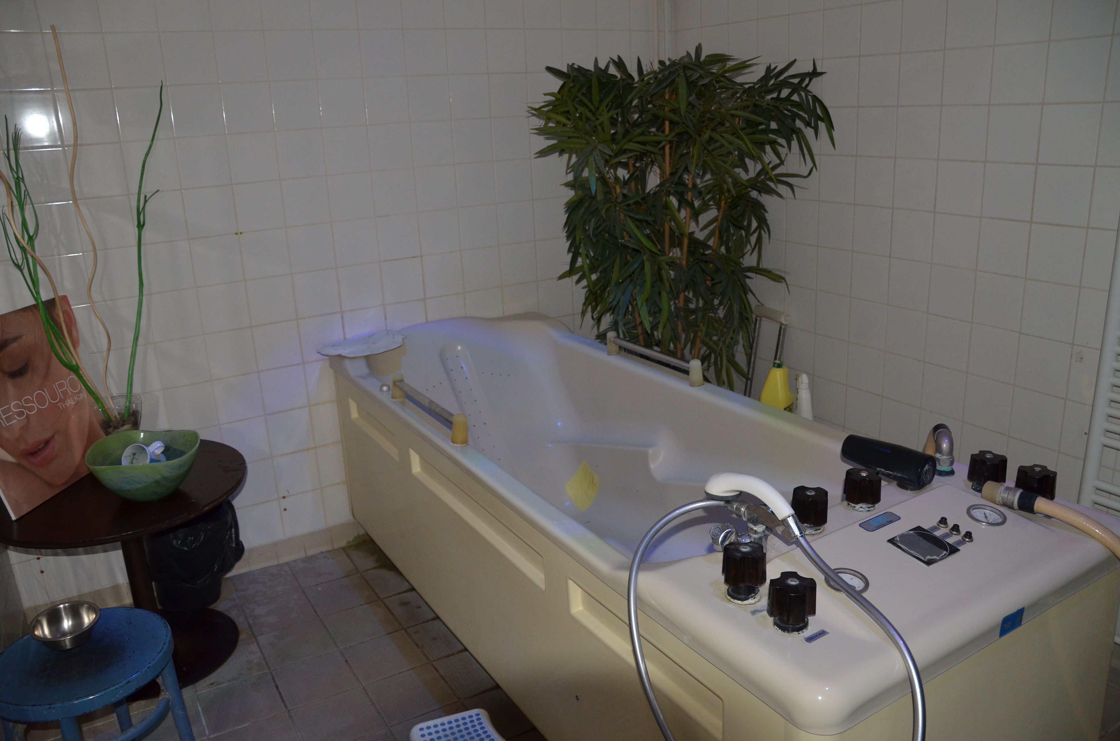 A bathroom with a bathtub, a green bowl on a table, a blue stool, and a potted plant.