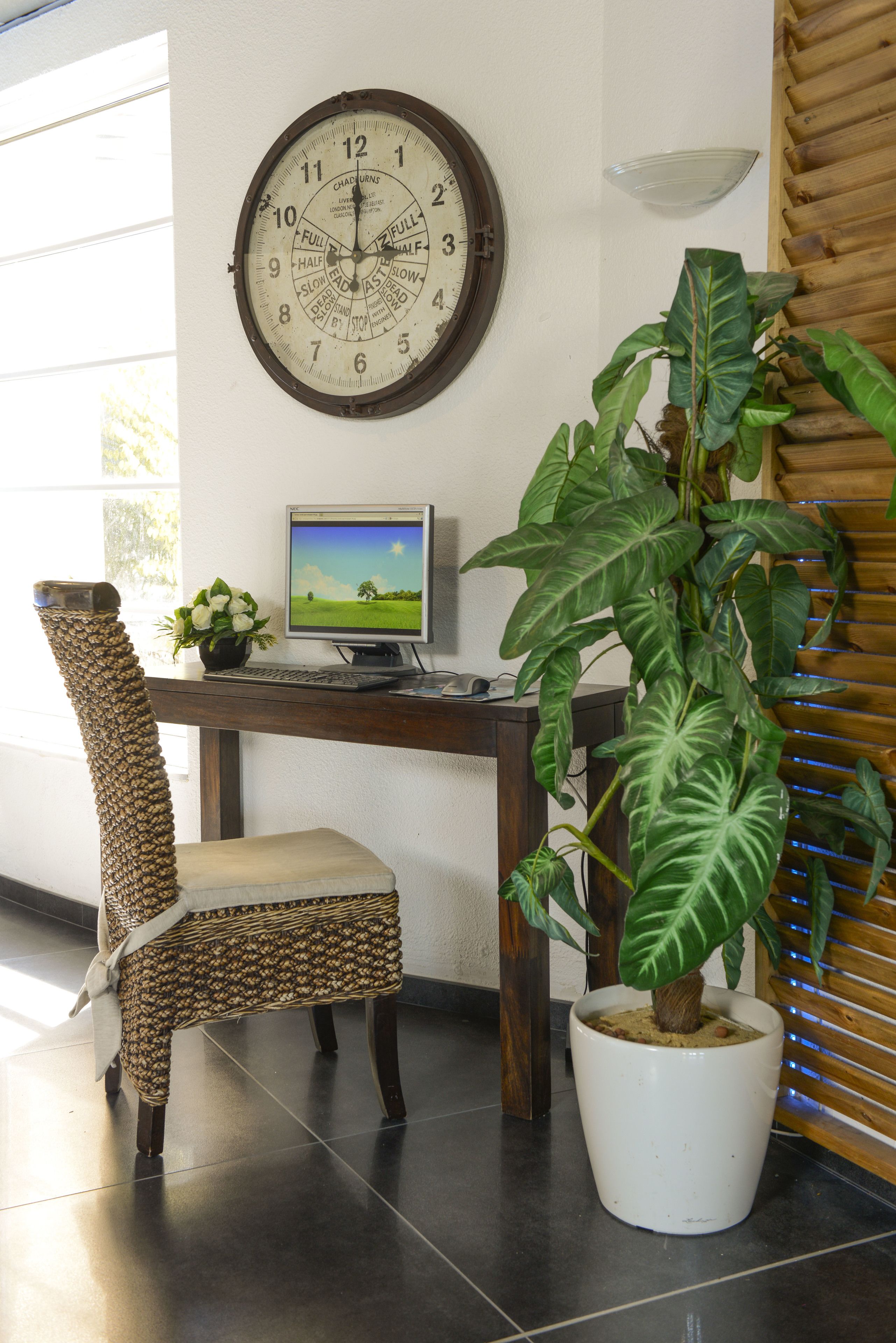 A room with a wooden desk, a computer, a chair, a large clock, and a potted plant.
