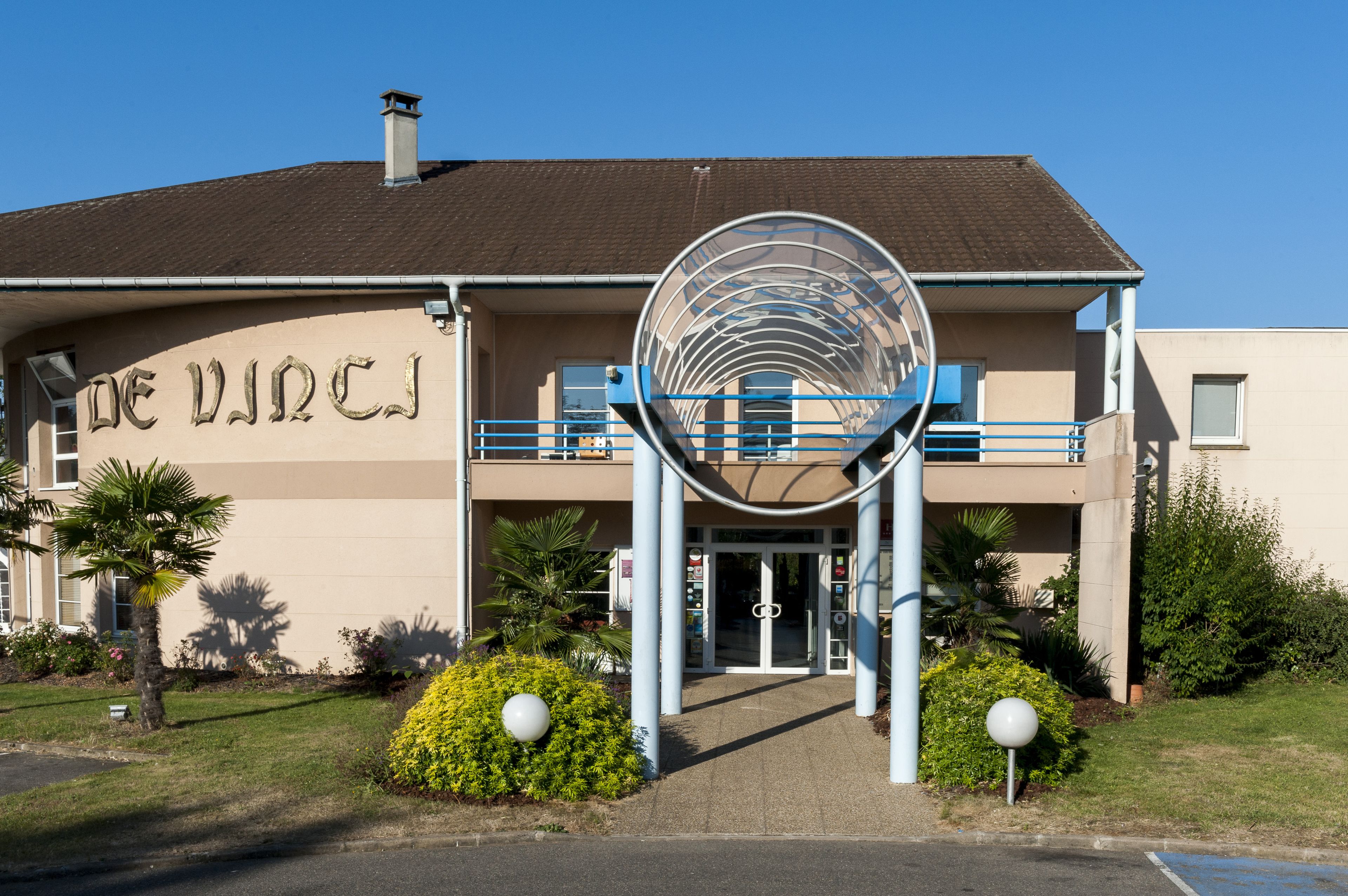 A building with a circular entrance structure, a sign reading "DE VINCJI", and a parking area with a blue line.
