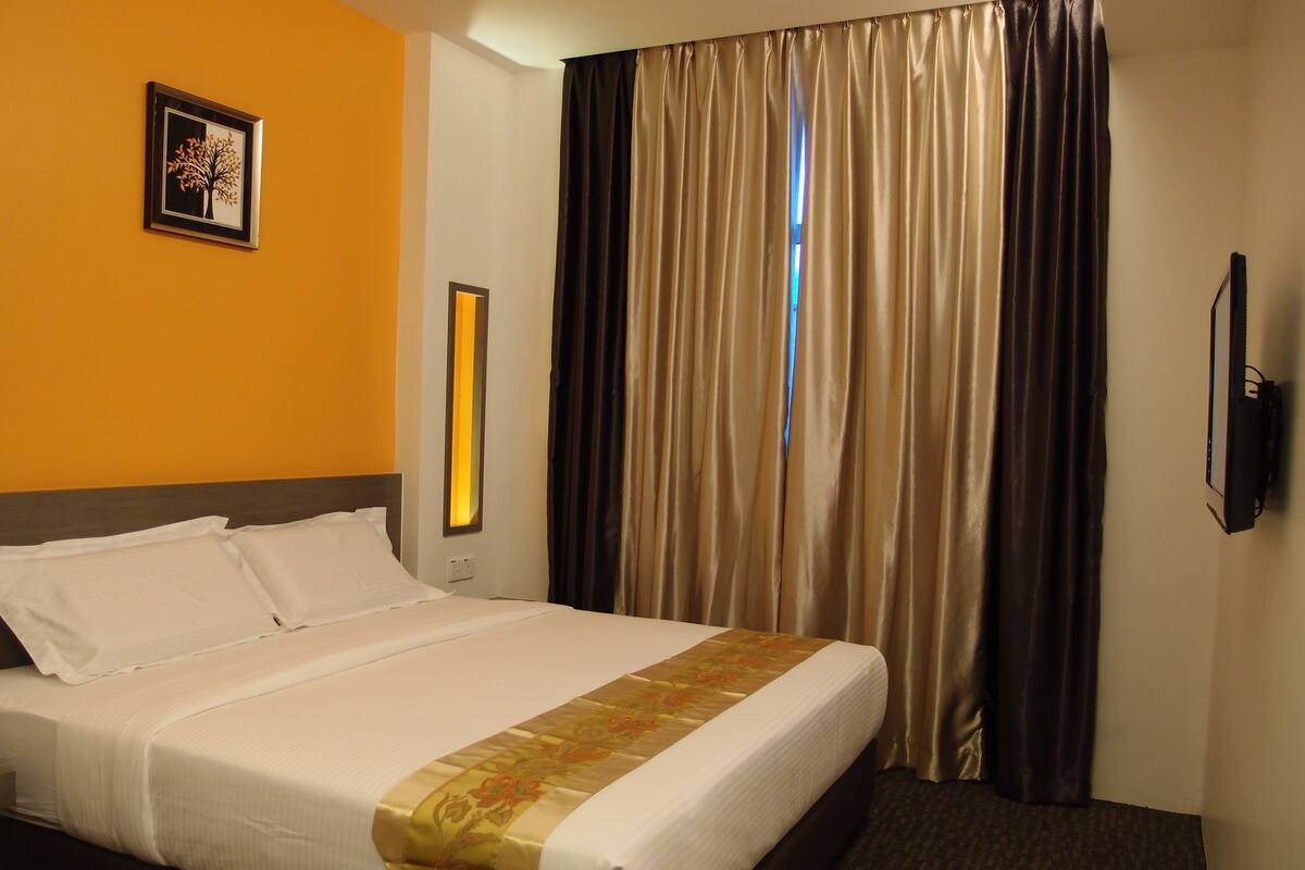 Ein Hotelzimmer mit einem Bett, einem gerahmten Bild an der Wand, einem an der Wand befestigten Fernseher und einem Fenster mit Vorhängen.