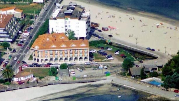 An aerial view of a coastal area with a large building with a red roof, a sandy beach, and a marina with boats.