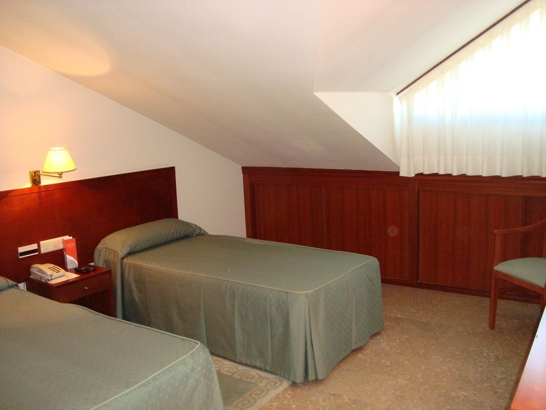 A hotel room with two beds, a wooden headboard, a nightstand with a lamp, and a window with curtains.