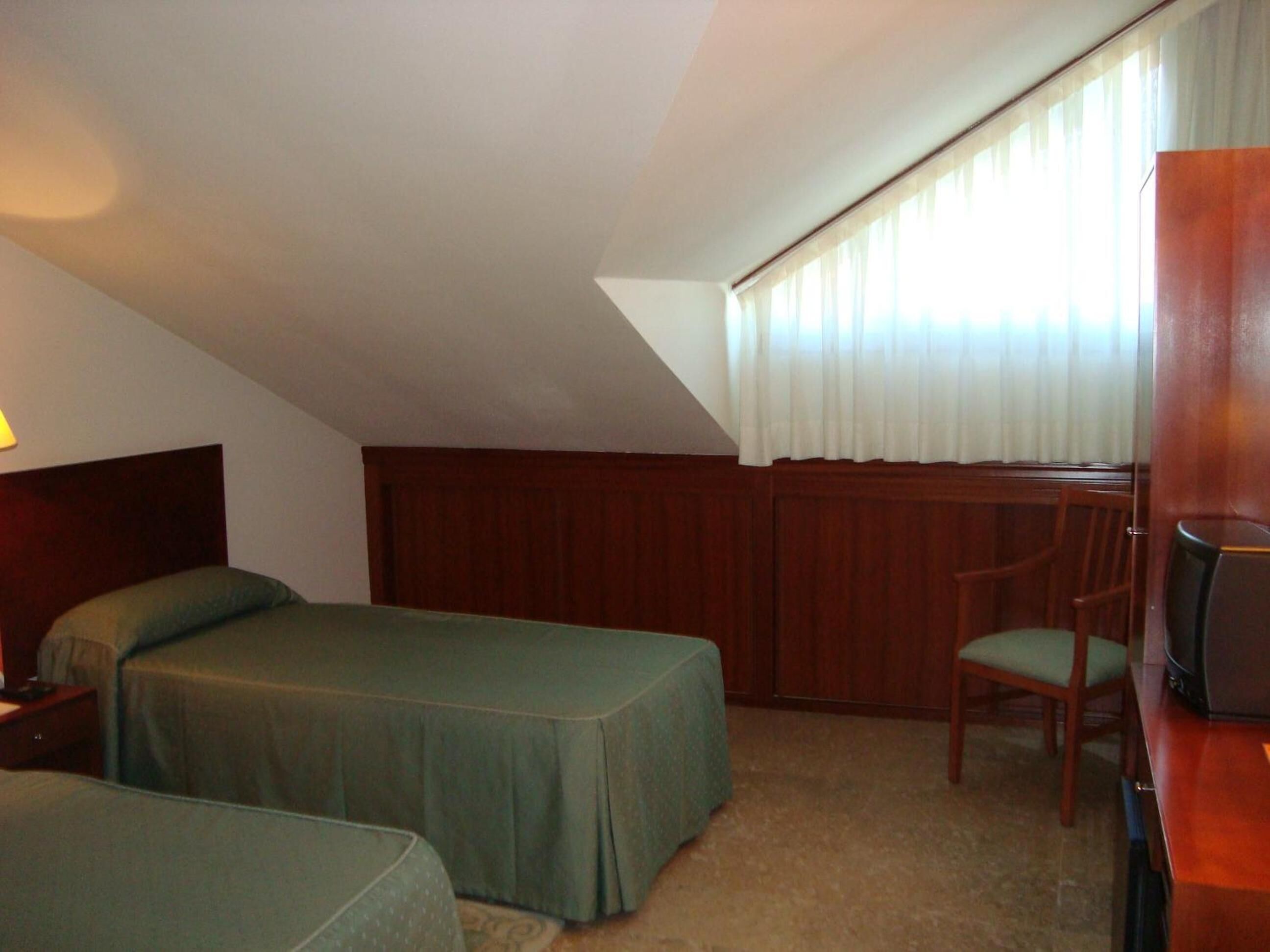 A room with two single beds, a wooden headboard, a chair, a desk with a computer, and a window with curtains.