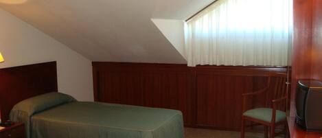 A room with two single beds, a wooden headboard, a chair, a desk with a computer, and a window with curtains.