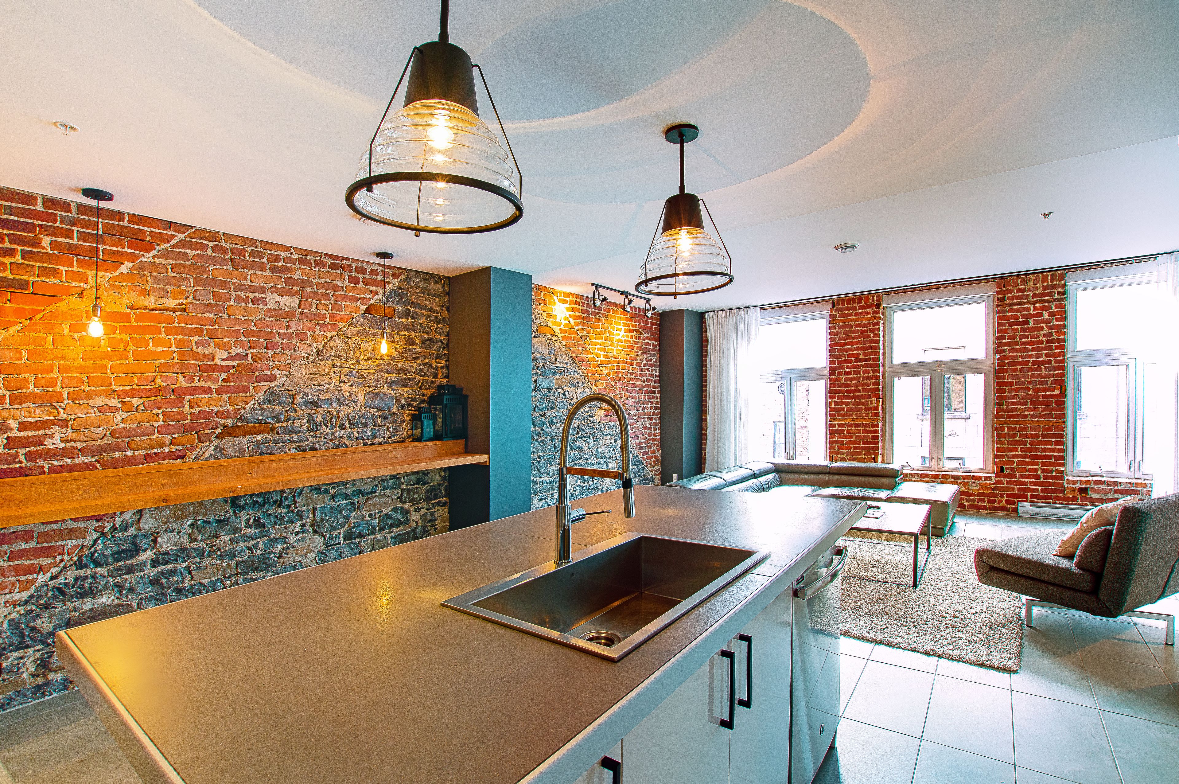 A modern kitchen with a large island, a brick accent wall, pendant lighting, and a view of the city through large windows.