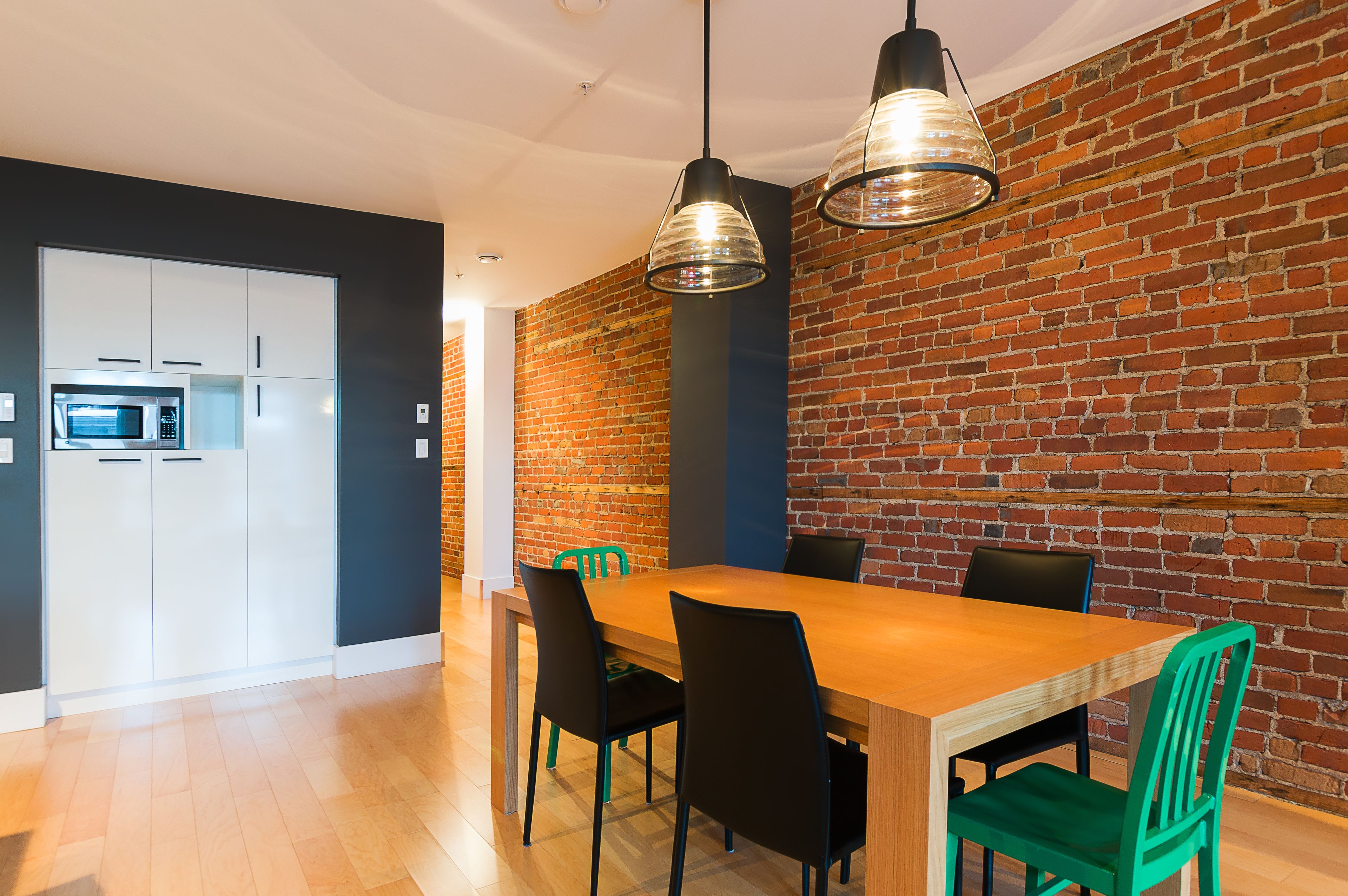 A modern kitchen with white cabinets, a microwave, and a brick wall. There is a wooden dining table with black and green chairs.