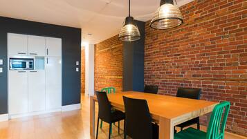 A modern kitchen with white cabinets, a microwave, and a brick wall. There is a wooden dining table with black and green chairs.