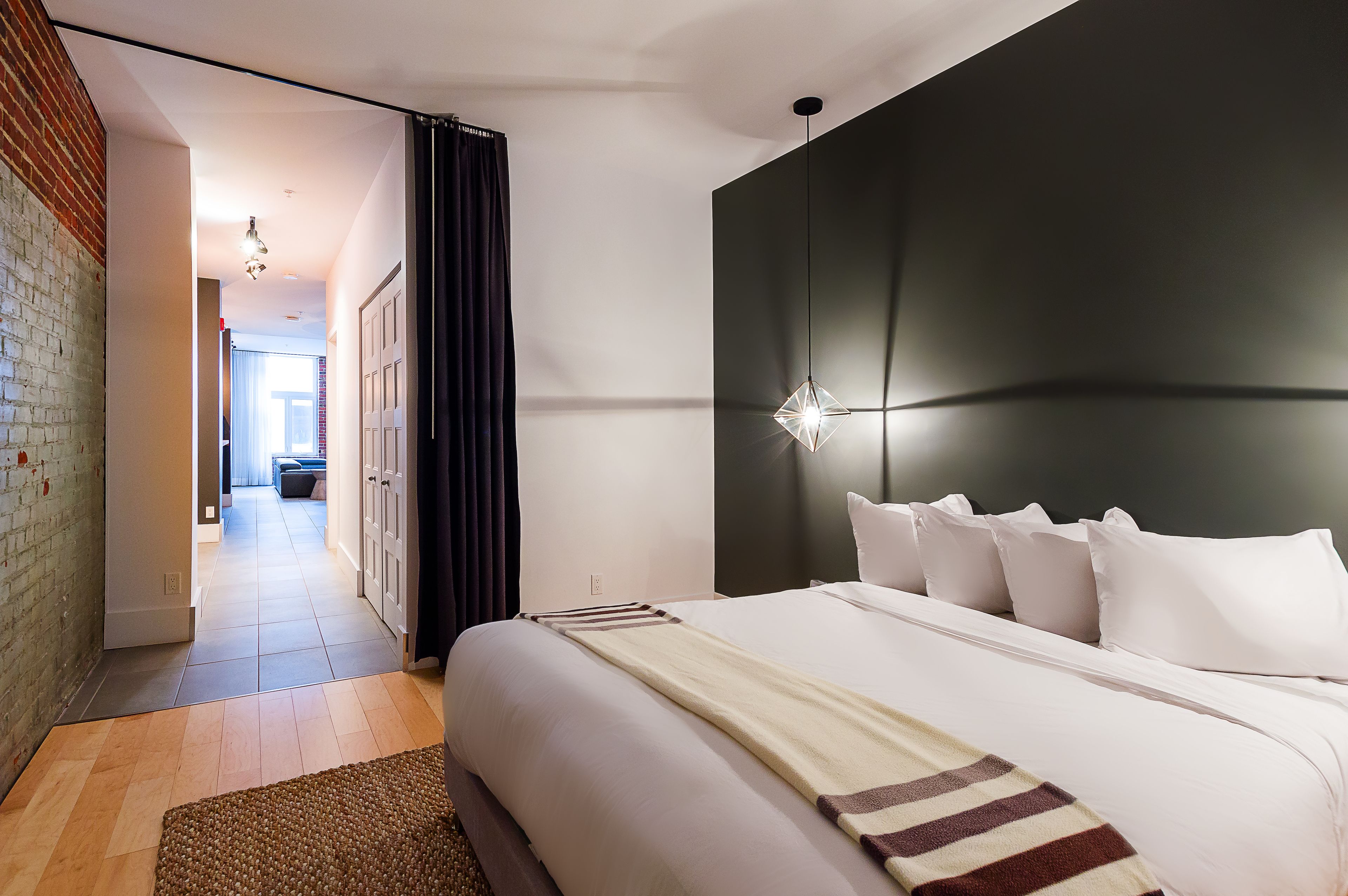 A modern hotel room with a large bed, a dark accent wall, and a hallway leading to another room.