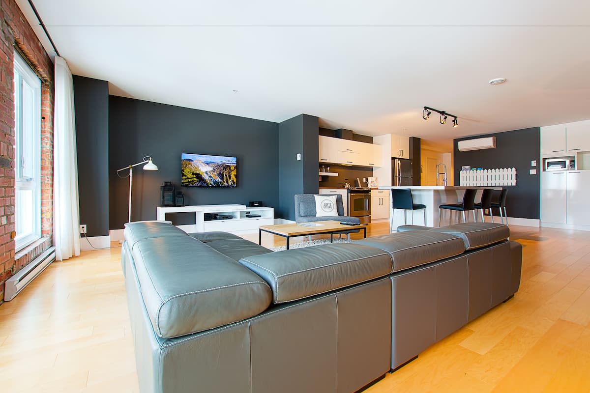 Una sala de estar moderna con un sofá seccional gris, un televisor de pantalla plana y una cocina con gabinetes blancos y un refrigerador de acero inoxidable.