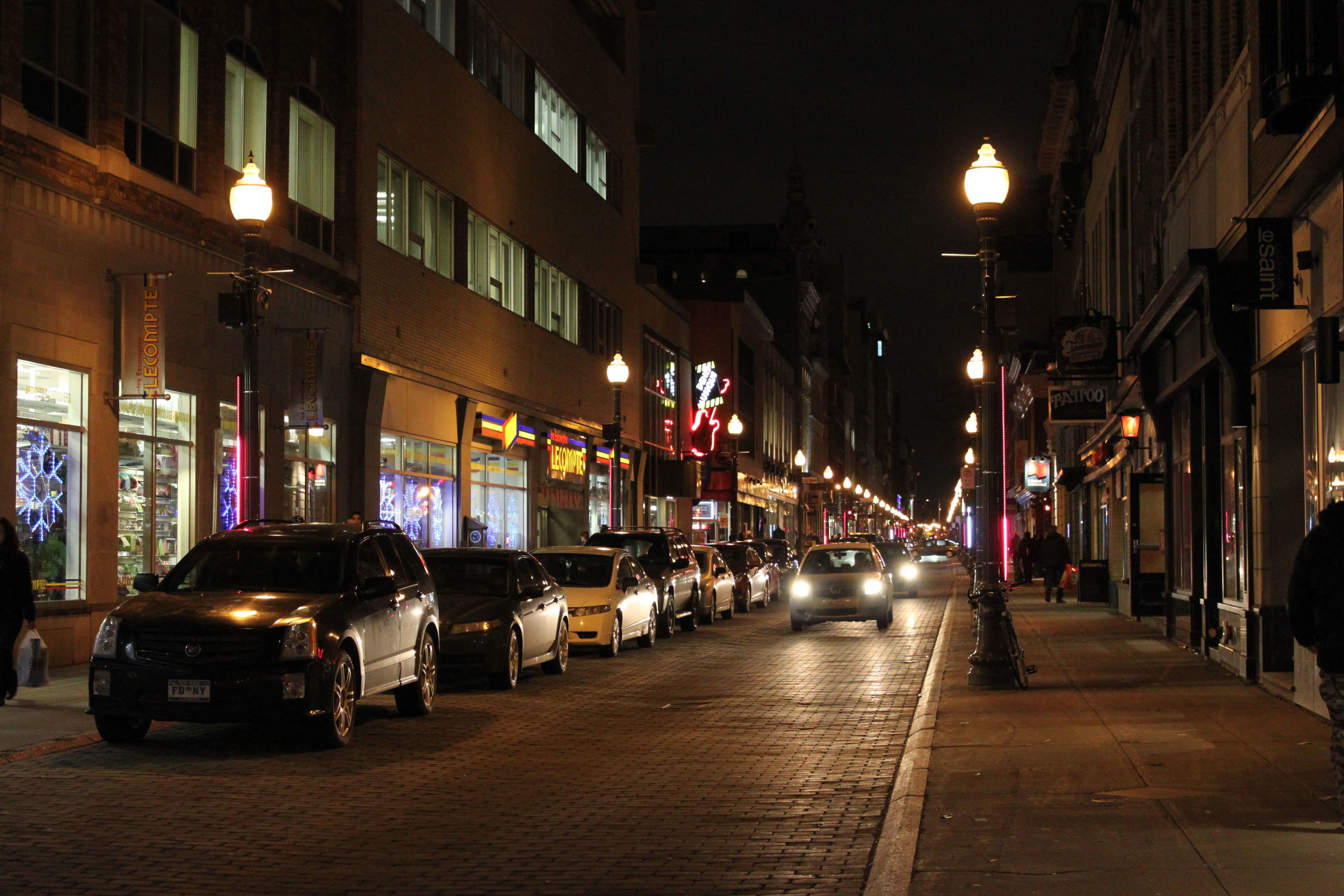 Une scène de rue nocturne, avec des voitures garées et des devantures de magasins illuminées.