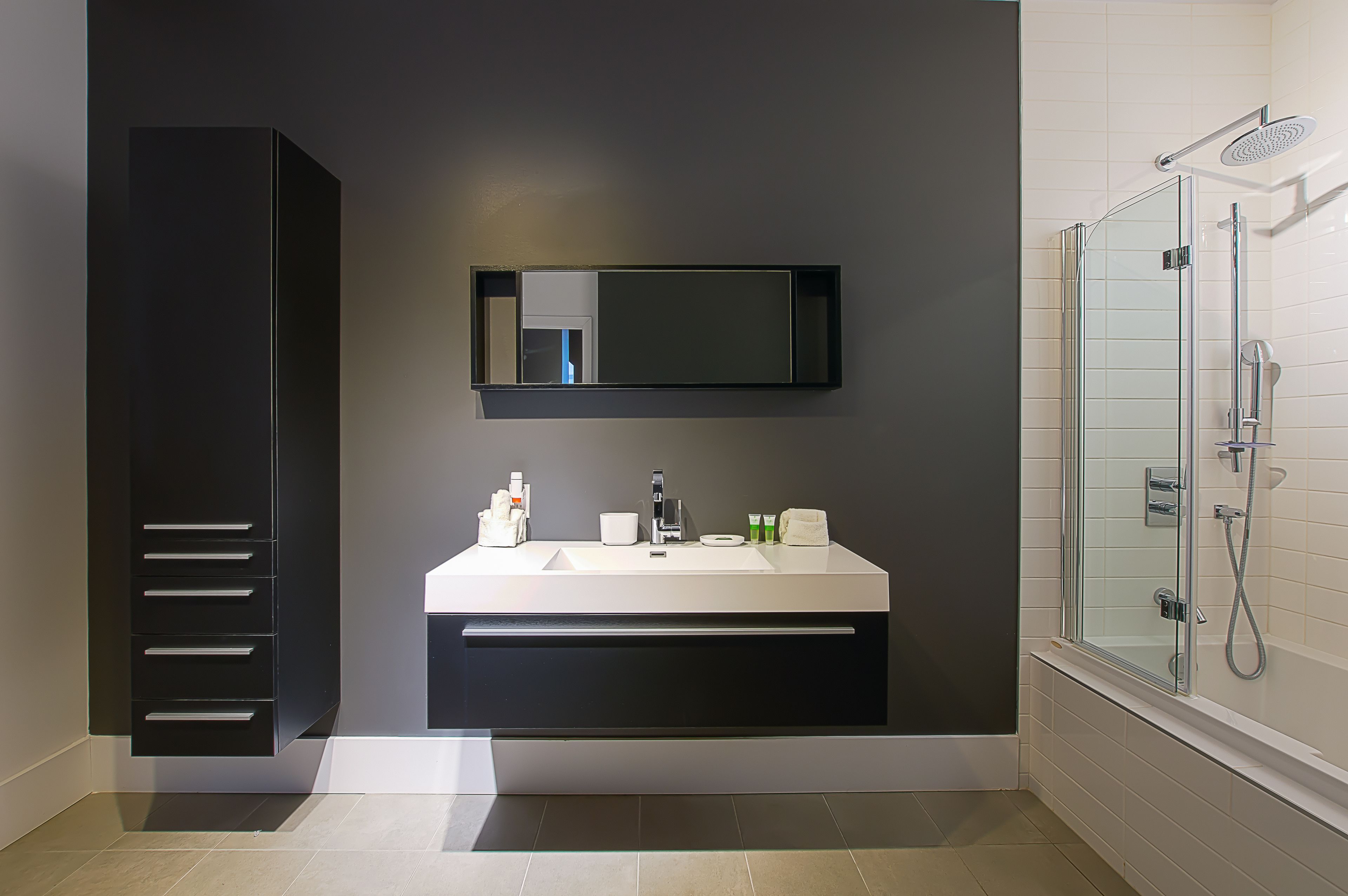 Een moderne badkamer met een donker kastje, een grote spiegel, een wit wastafel en een douche met glazen wanden.