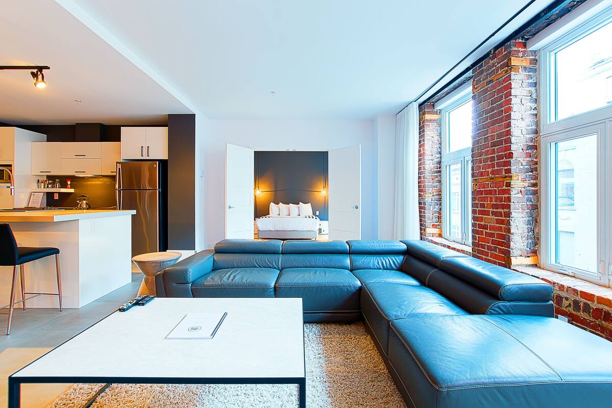 Un espacio moderno para vivir con cocina, un amplio sofá de cuero azul en forma de L y vistas a un muro de ladrillo a través de amplias ventanas.