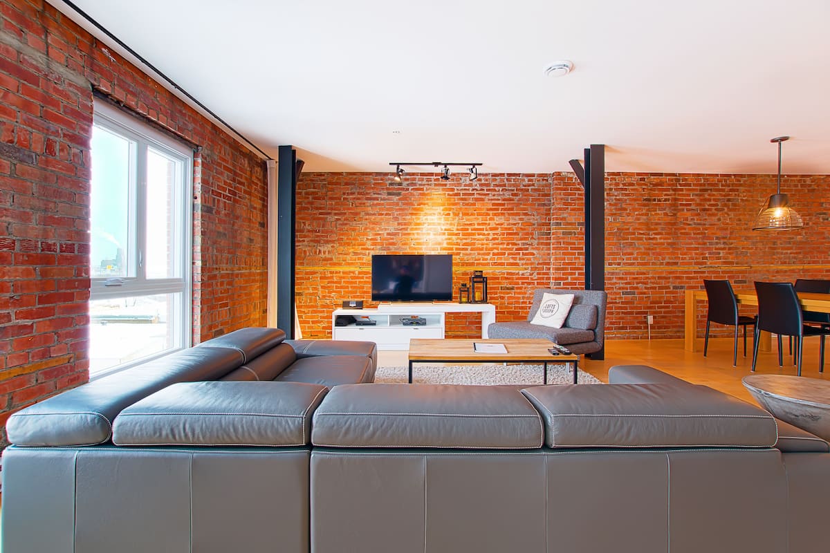 Una sala de estar moderna con pared de ladrillo, un sofá de cuero grande, un televisor de pantalla plana y una zona de comedor con sillas y una mesa.