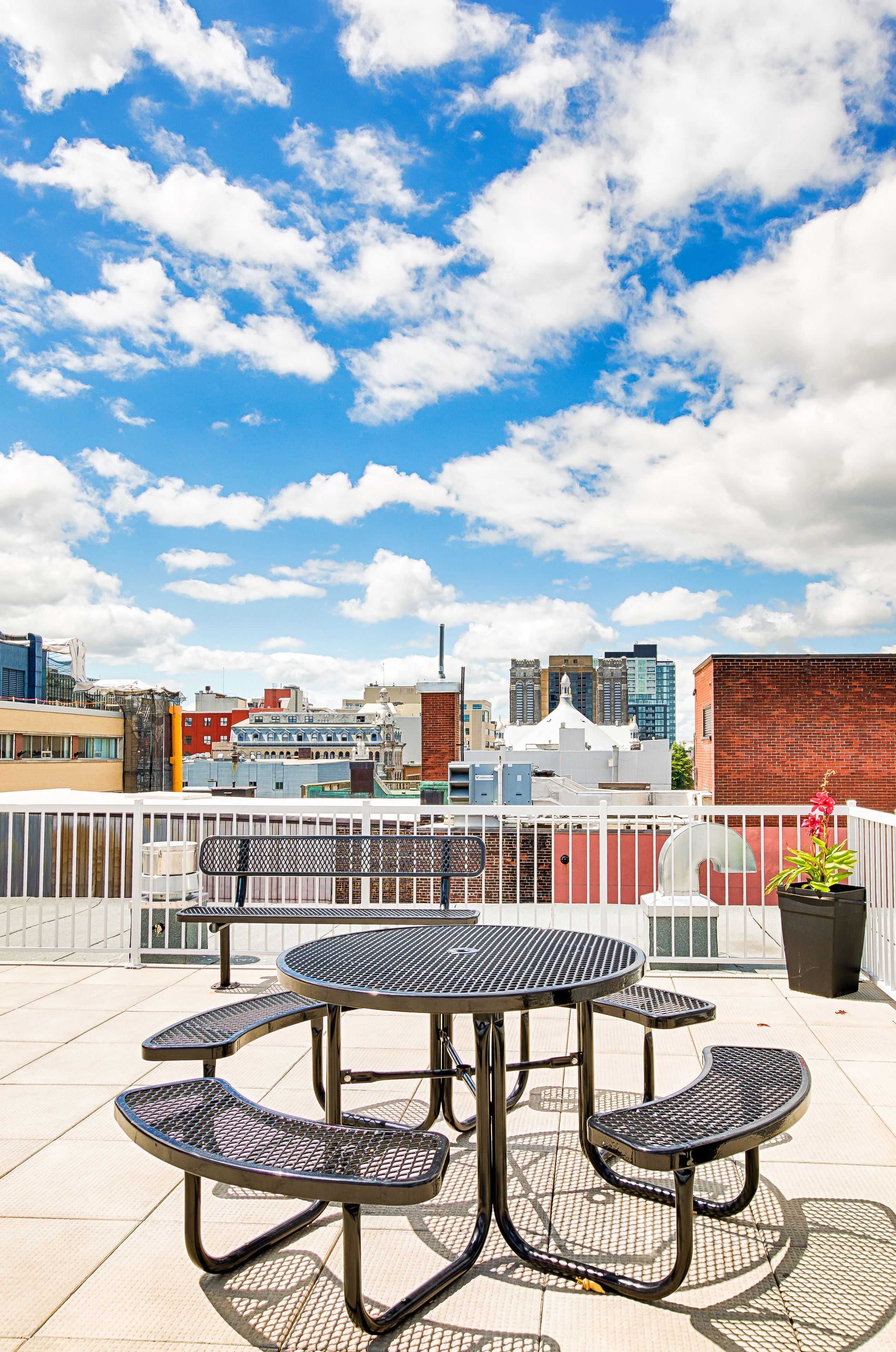 Um terraço no telhado com uma mesa redonda e bancos, com vista para um horizonte urbano com diversos edifícios.