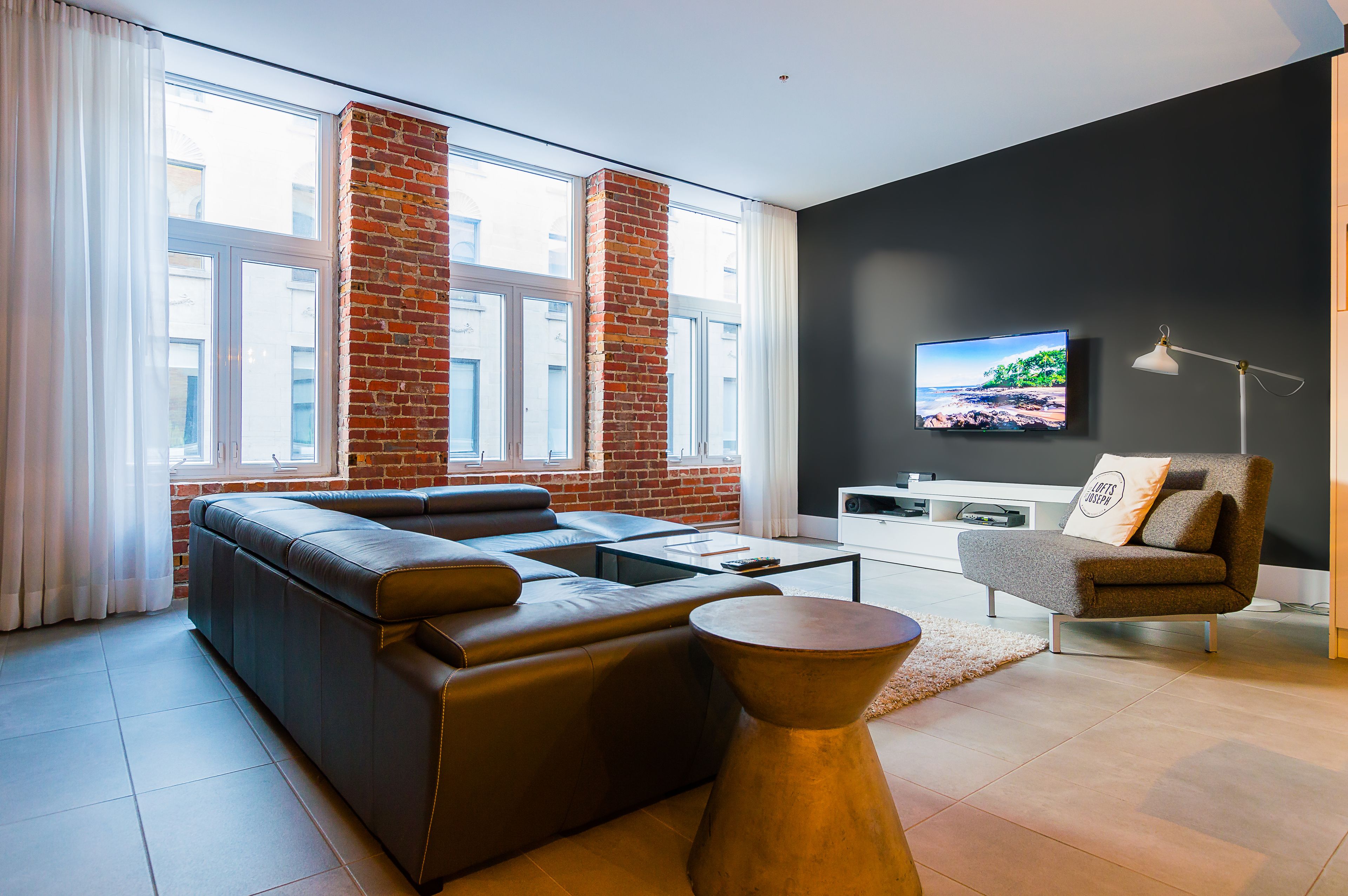 Una sala de estar moderna con un sofá de cuero, una mesa de centro, una mesita auxiliar, un televisor montado en la pared y una pared de ladrillo decorativa.