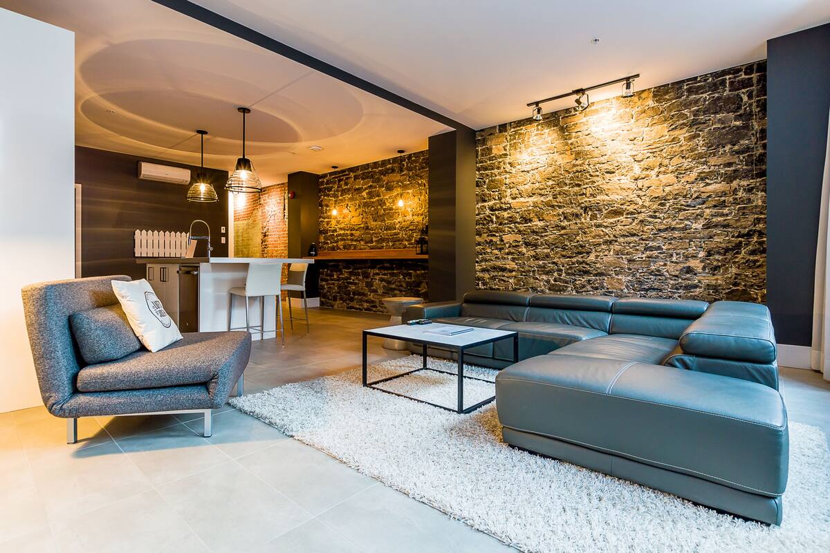 Una sala moderna con un sofá modular, un sillón gris y una mesa de centro. Se observa una pared de piedra y una valla blanca al fondo.