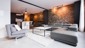 Een moderne woonkamer met een hoekbank, een grijze fauteuil en een salontafel. Achtergrond is een bakstenen muur en een trap.