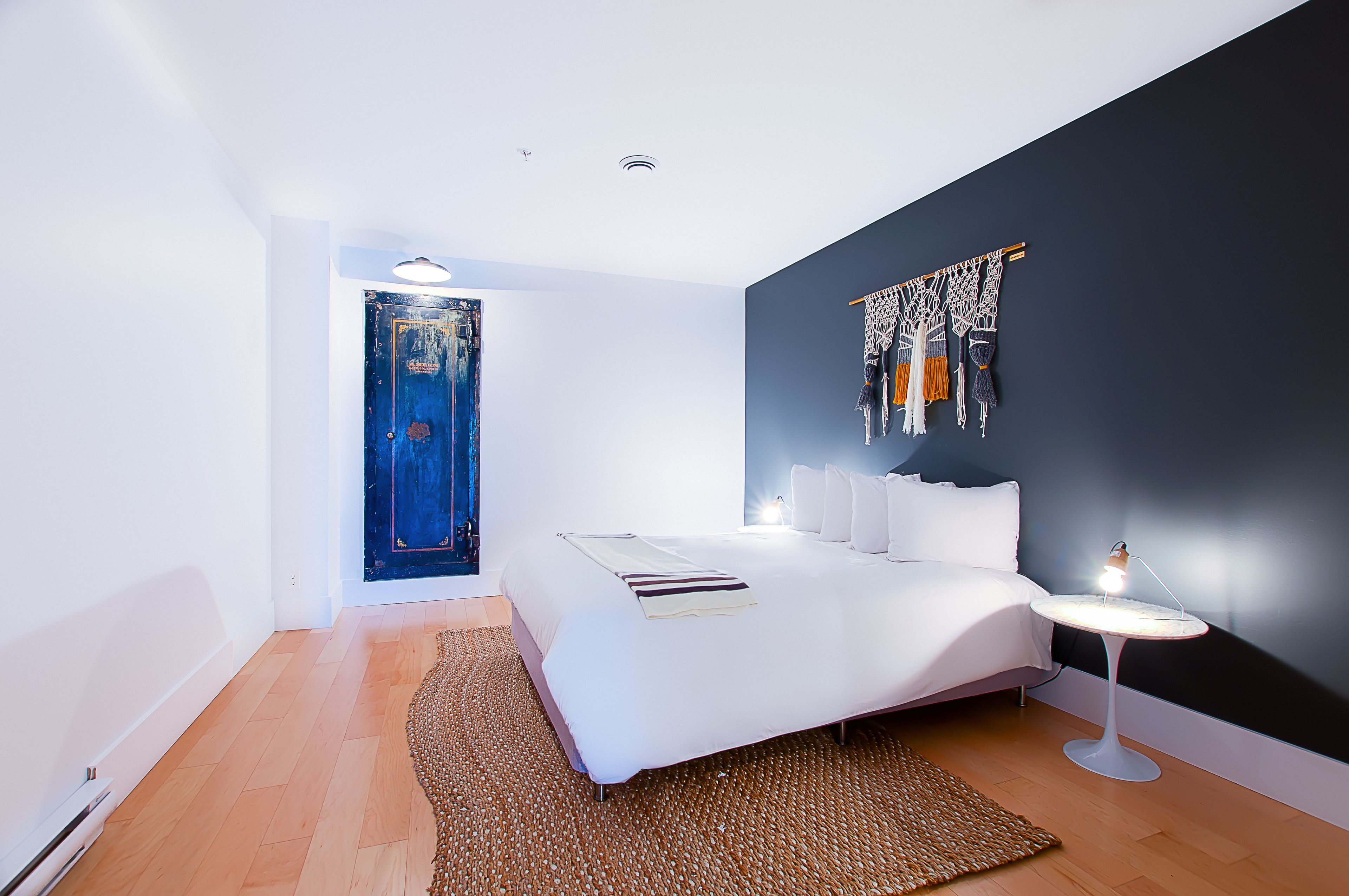 Un dormitorio moderno con una cama grande, una mesita de noche con lámpara, una pared decorativa oscura y una puerta azul única.