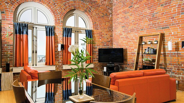 A living room with a brick wall, orange sofa, and a flat-screen TV. - Les Lofts 1048 (Québec City)