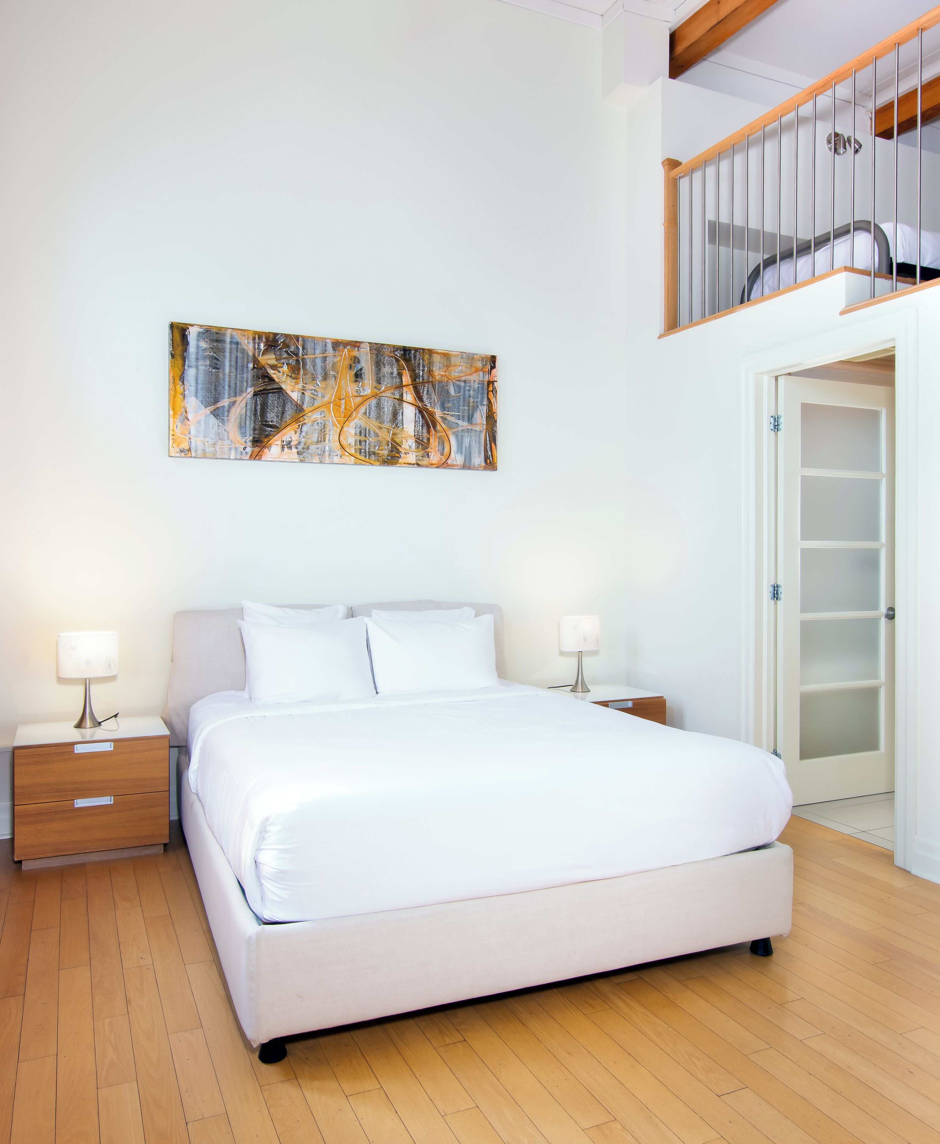 A bedroom with a large bed, two bedside tables with lamps, a wooden floor, a white door, and a loft space above.