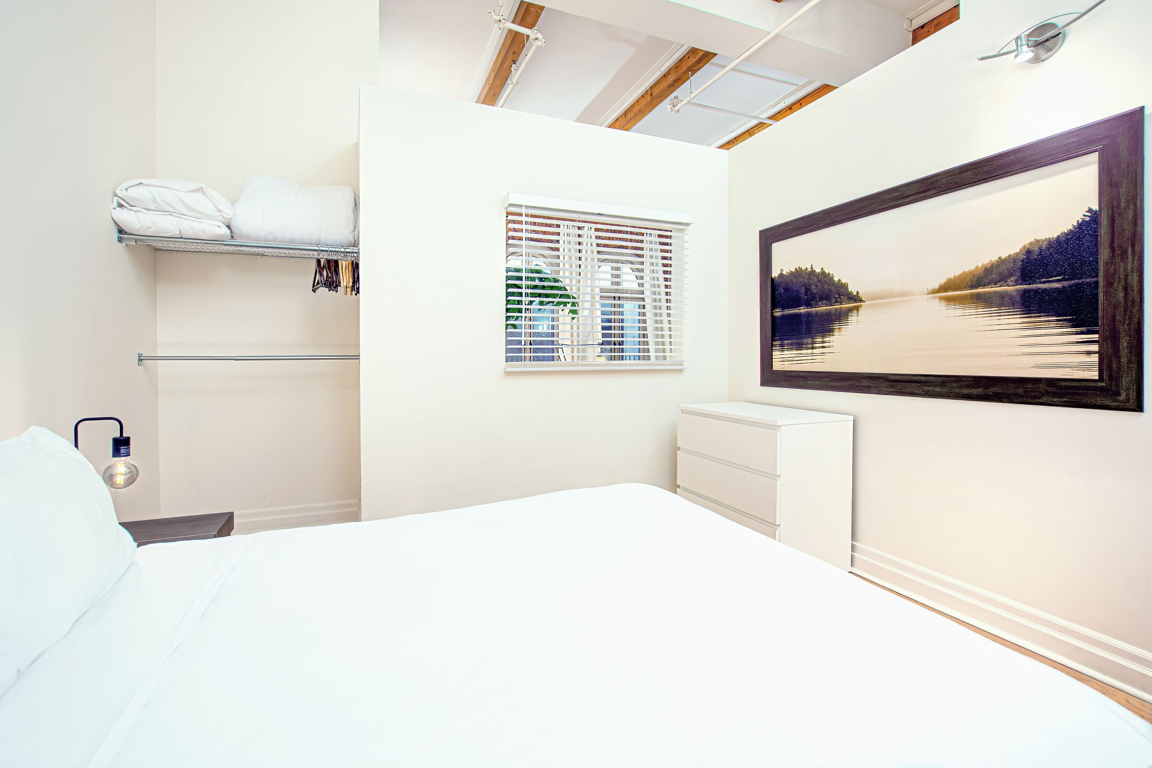 A bedroom with a bed, a window with blinds, a wall-mounted shelf with towels, and a framed picture on the wall.