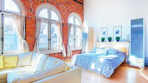 A bedroom with a brick wall, two beds, a sofa, and large windows. - Les Lofts 1048 (Québec City)