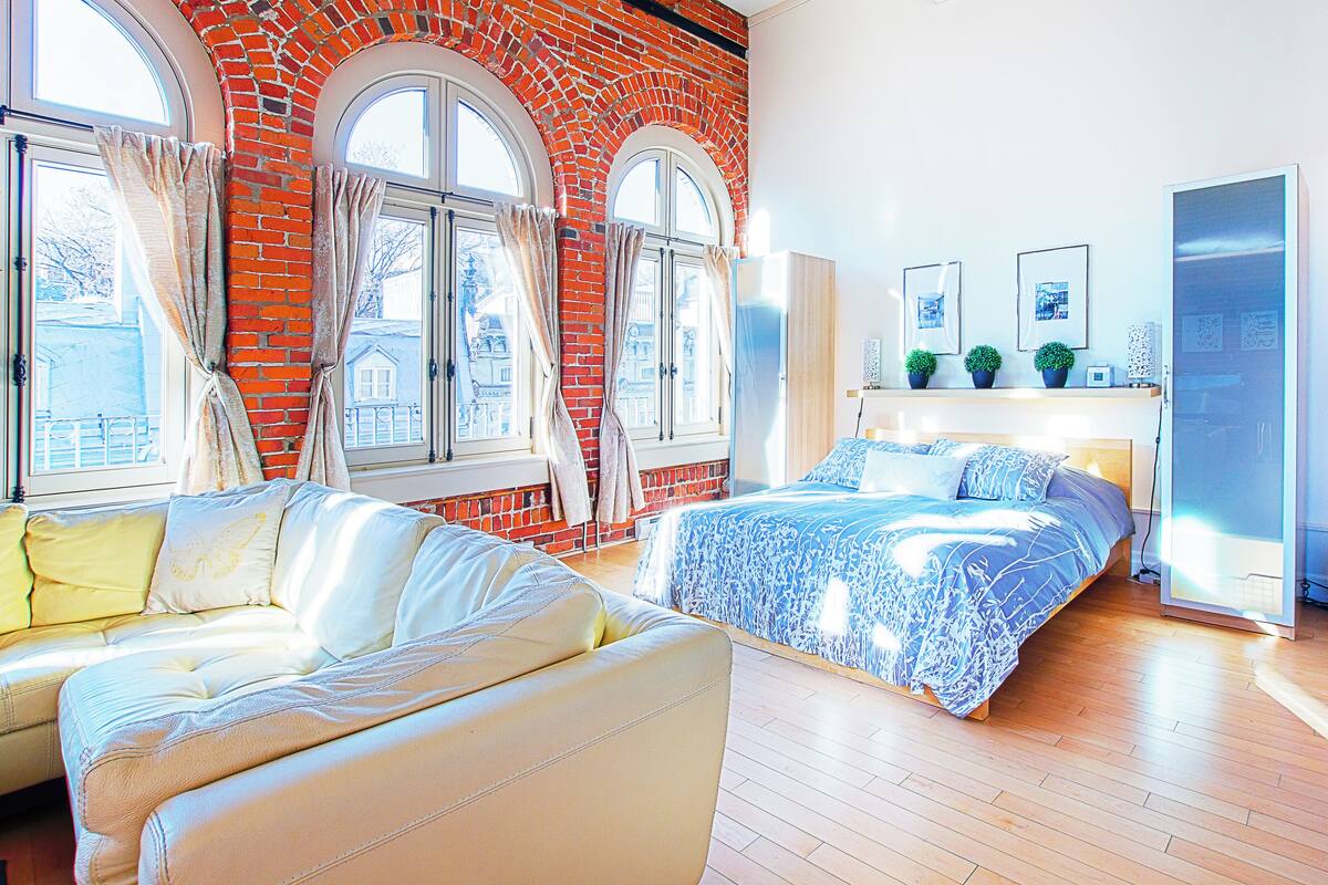 A bedroom with a brick wall, two beds, a sofa, and large windows.