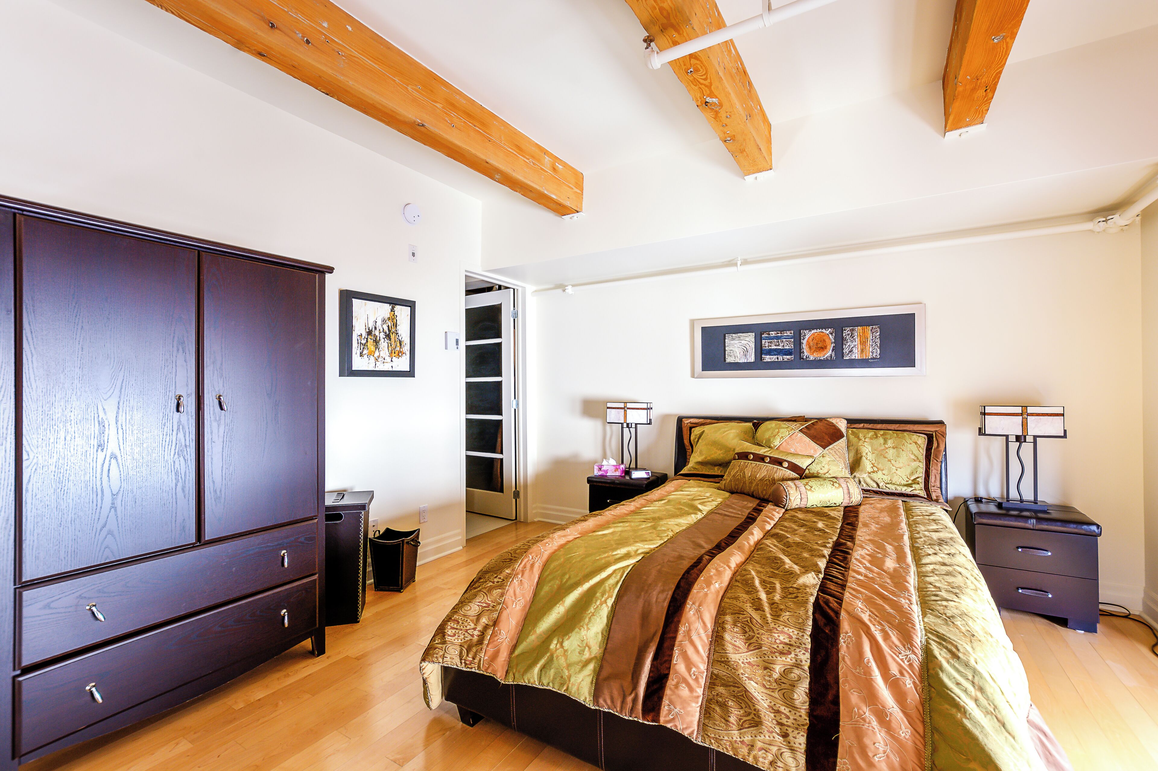 A bedroom with a large wardrobe, a bed with patterned bedding, a nightstand, and a wall-mounted lamp.