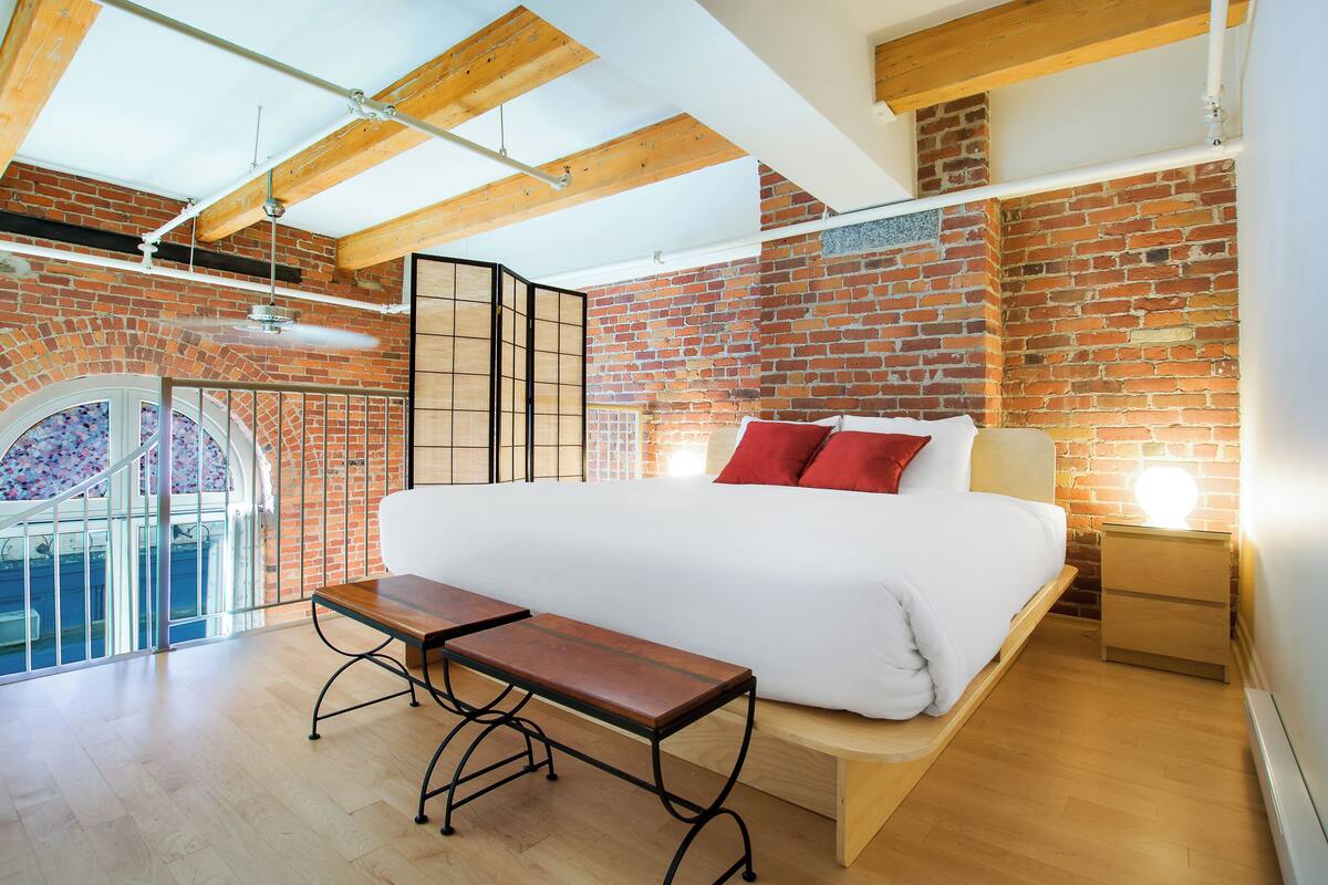 A bedroom with a large bed, wooden bench, brick walls, and a skylight.