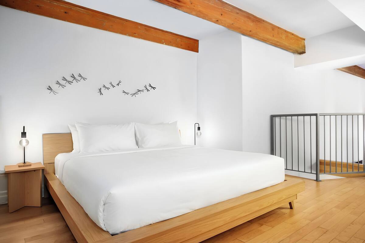 A modern bedroom with a wooden bed frame, white bedding, and a nightstand with a lamp.