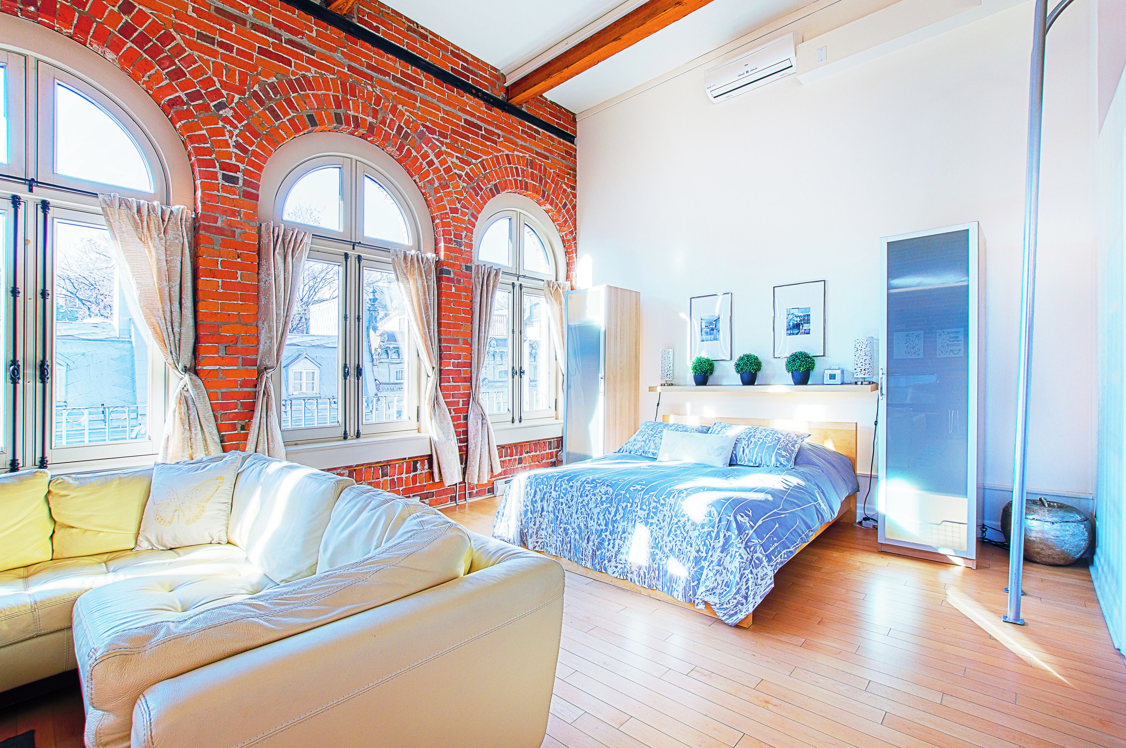 A bedroom with a brick wall, large windows, a bed, a sofa, and a wardrobe.
